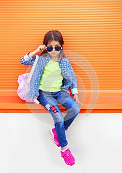 Fashion little girl child wearing a sunglasses, shirt, jeans and backpack over colorful orange