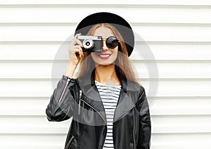 Fashion look, pretty cool young woman model with retro film camera wearing elegant hat, leather rock jacket over white