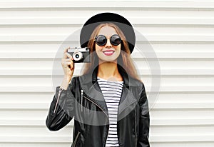 Fashion look, pretty young woman model with retro film camera wearing elegant black hat, leather rock jacket over white