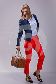 Fashion look of woman dressed red pants, blue jacket, brown bag. model looks at a leather bag. full height