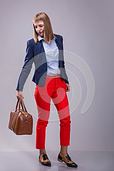 Fashion look of woman dressed red pants, blue jacket, brown bag. model looks at a leather bag. full height