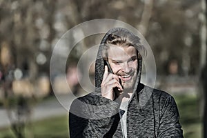 Fashion macho smiling with smartphone in casual sweatshirt. Happy guy in hood talk on mobile phone on sunny outdoor. Bearded man s