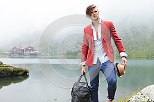 Fashion man holding his hat and travel bag near lake