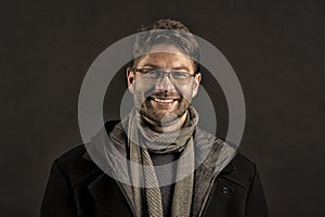 Fashion model with beard smile on dark background. Happy man in glasses on bearded face. Bearded man in scarf and coat