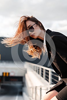 Fashion model beautiful young girl in dark vintage sunglasses in black stylish blazer in summer windy sunny day outdoors. Beauty