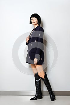 Fashion model with bob haircut posing near the wall in blue jacket with gems decor and in wide high boots