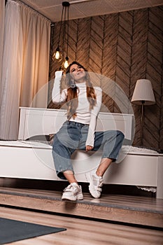 Fashion model of a chic young woman in a white stylish top in fashionable blue jeans posing on the bed in the bedroom. Red-haired