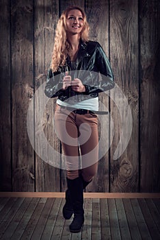 Fashion model with curly hair dressed in black jacket, denim pants and tall boots over wooden wall background
