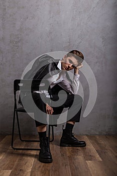 Fashion model of an elegant young man in a black leather jacket in a classic white shirt in trendy boots sits on a chair near a