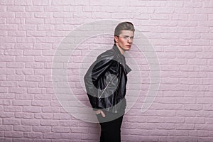 Fashion model of a European stylish young man in a fashionable black leather jacket with a trendy hairstyle stands near a brick