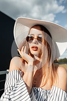 Fashion model lovely young woman in fashionable shirt in stylish sunglasses in vintage straw hat rests outdoors on bright sunny