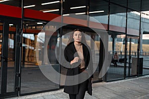 Fashion model pretty stylish young woman in a fashionable black coat in a beige shirt walks near a vintage glass building in the