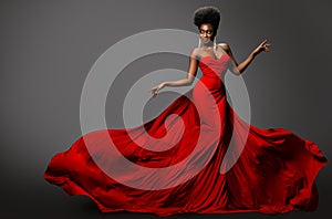 Fashion Model in Red Long waving Luxury Dress. Dark Skinned Beauty Woman with Afro Black Hairstyle dancing over Gray Background.