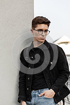 Fashion model serious young man in stylish jeans youth casual wear in fashionable glasses with backpack rest near wall in city.