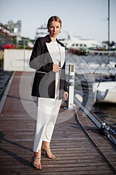 Fashion model strikes a pose on a pier near water, adorned in luxurious attire long white dress and a black blazer