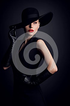 Fashion Model in Wide Brim Hat with Red Lips Make up. Elegant Retro Woman in Black Suit Dress and Gloves over Dark Gray Studio photo