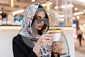 Fashion model young beautiful woman in stylish silk shawl in trendy sunglasses in black fashionable coat with cup of coffee is