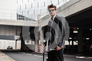 Fashion model young modern man in vintage sunglasses in a leather black stylish jacket in jeans stands on parking lot outdoors.