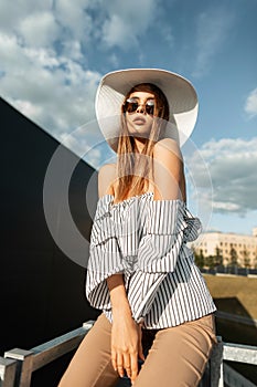 Fashion model young woman in fashionable shirt in stylish sunglasses in vintage straw hat in pants rests outdoors on bright sunny