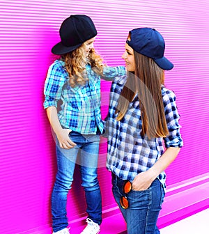 Fashion mother looking at child little girl in checkered shirts and baseball caps in city on colorful pink wall