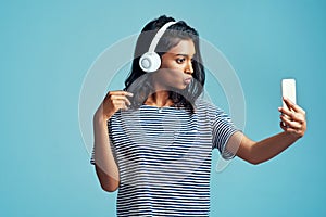 Fashion, music and selfie of woman in studio for social media isolated on blue background. Photography, headphones and