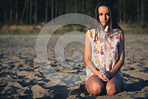 Fashion outdoor photo of beautiful sexy woman sitting on the sand with dark hair in light shirt