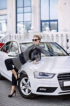 Fashion outdoor photo of beautiful woman with dark hair in black leather jacket and sunglasses posing in luxurious auto