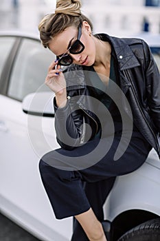 Fashion outdoor photo of beautiful woman with dark hair in black leather jacket and sunglasses posing in luxurious auto