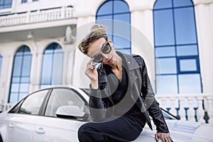 Fashion outdoor photo of beautiful woman with dark hair in black leather jacket and sunglasses posing in luxurious auto