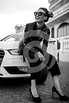 Fashion outdoor photo of beautiful woman with dark hair in black leather jacket and sunglasses posing in luxurious auto