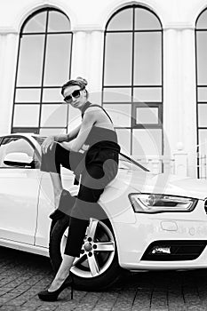 Fashion outdoor photo of beautiful woman with dark hair in black leather jacket and sunglasses posing in luxurious auto