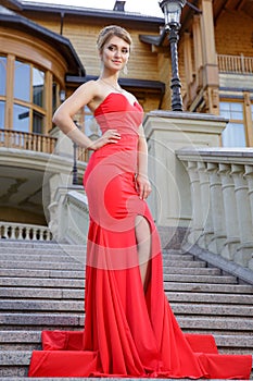 Fashion outdoor photo of beautiful woman in luxurious red dress posing on stairs in villa