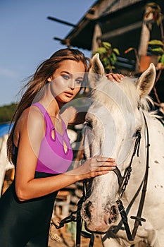 Beautiful young woman with dark hair in sportive clothes posing with white horse in stable