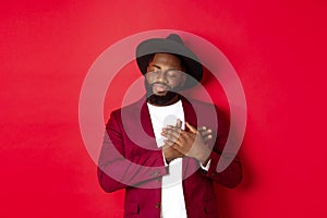 Fashion and party concept. Young bearded Black man in classy hat and jacket, holding hands on heart and smiling