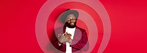 Fashion and party concept. Young bearded Black man in classy hat and jacket, holding hands on heart and smiling