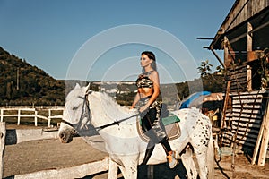 Beautiful woman with dark hair in elegant sportive suit posing near white horse in stable