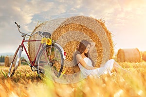 Fashion photo, beautiful woman sitting in front of bales of wheat, next to the old bike