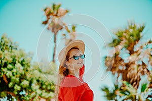 Fashion photo of a girl in black glasses with a chain and a large straw hat, a red dress on a background of palm trees. Wind in