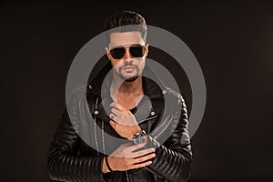 Fashion photo of handsome man wearing black leather jacket, sunglasses, hand watch, posing on dark studio background