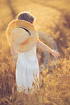 Fashion photo of a little girl in white dress and straw hat at the evening wheat field