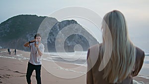 Fashion photographer taking picture at ocean shore. Blonde model posing rear