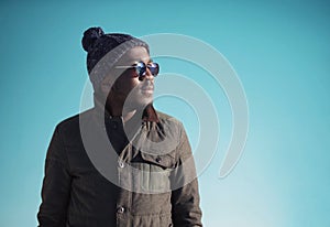 Fashion portrait african man wearing sunglasses, jacket, knitted hat in winter day over blue sky background