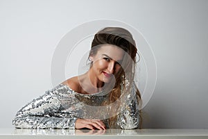 Fashion portrait of a beautiful smiling woman with long hair in a pretty silver dress with tinsel sitting at the table