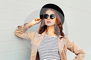 Fashion portrait beautiful young woman with red lips wearing black hat sunglasses coat over grey background