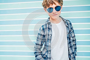 Fashion portrait of caucasian blonde hair 12 year old teenager boy dressed white t-shirt with checkered shirt in blue sunglasses