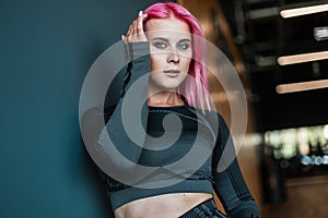 Fashion portrait of a fitness girl with pink hair in black sportswear on a dark background in the gym