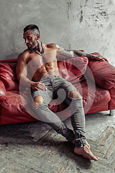 Fashion portrait of naked male model with tattoo and a black beard sitting in hot pose on red leather sofa. Loft room interio