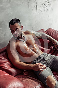 Fashion portrait of naked male model with tattoo and a black beard sitting in hot pose on red leather sofa. Loft room interio