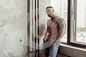 Fashion portrait of naked male model with tattoo and a black beard standing in hot pose on near the window. Loft room interio