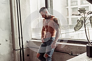 Fashion portrait of naked male model with tattoo and a black beard standing in hot pose on near the window. Loft room interio